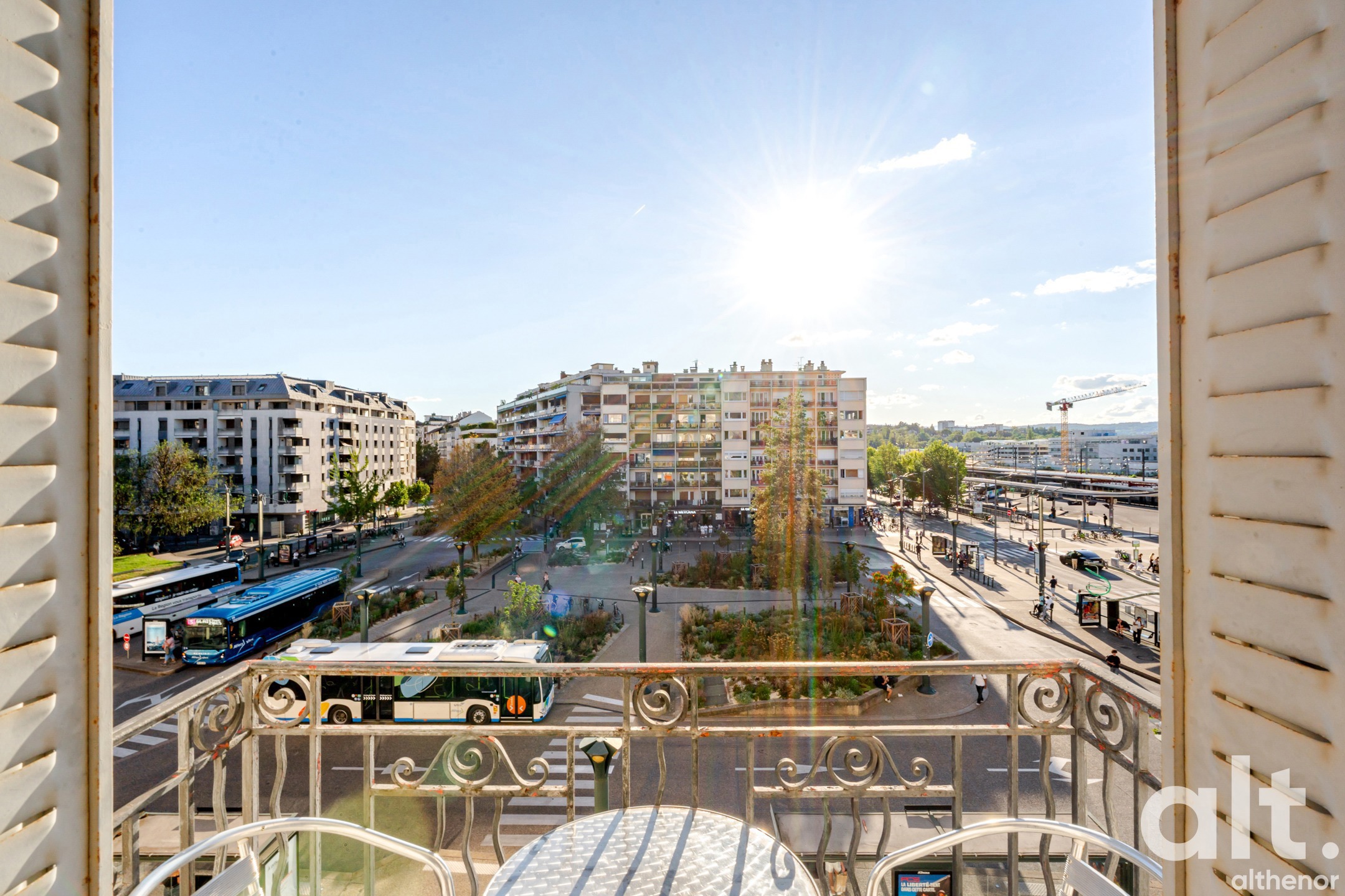 Balcony with a view of an urban square surrounded by residential buildings, white garden furniture, ornate gilt railing, clear sky.