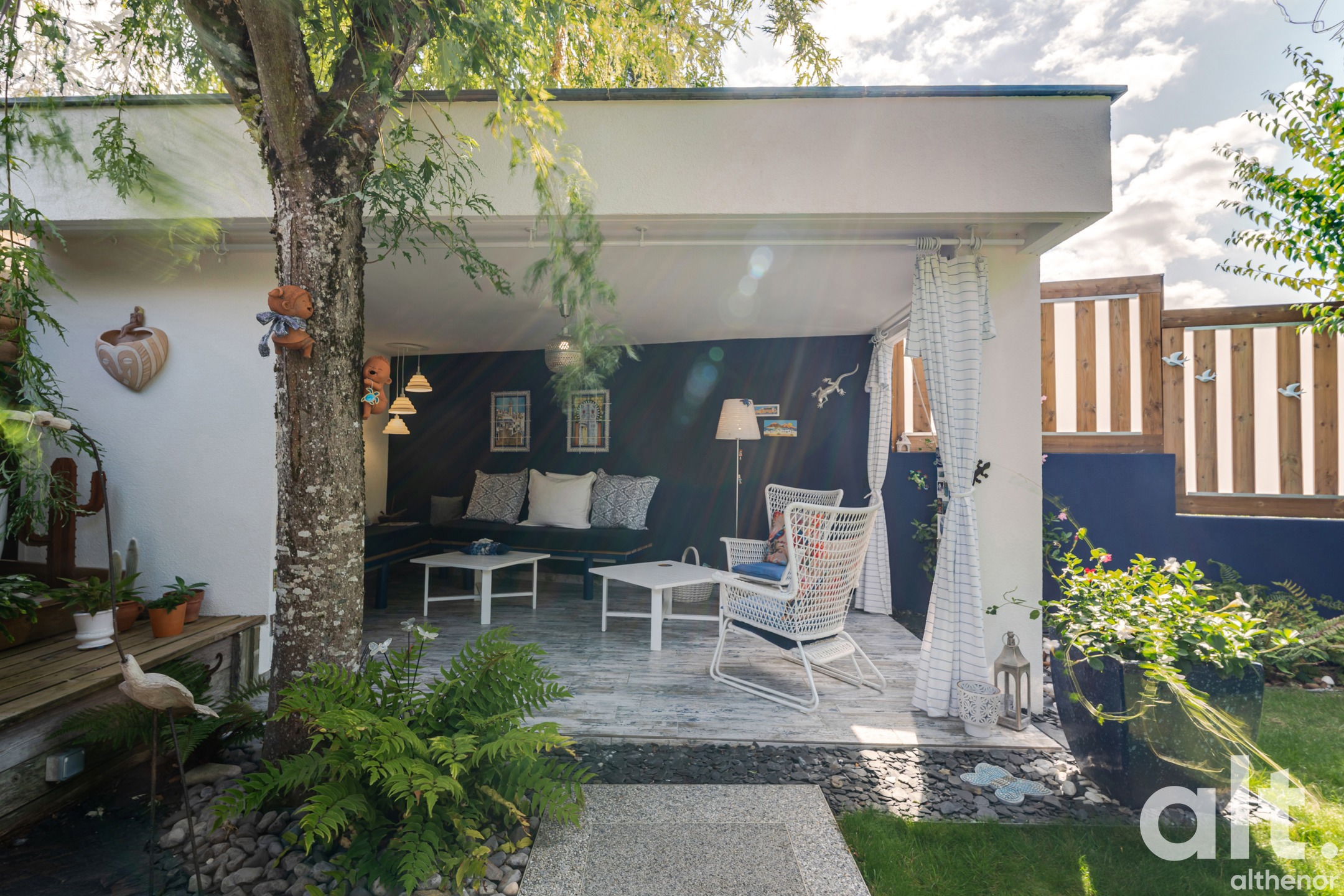 Exterior, covered terrace with a navy blue sofa, white armchair, coffee tables, under a white awning. Large tree, green plants, access to a side garden with a wooden fence.