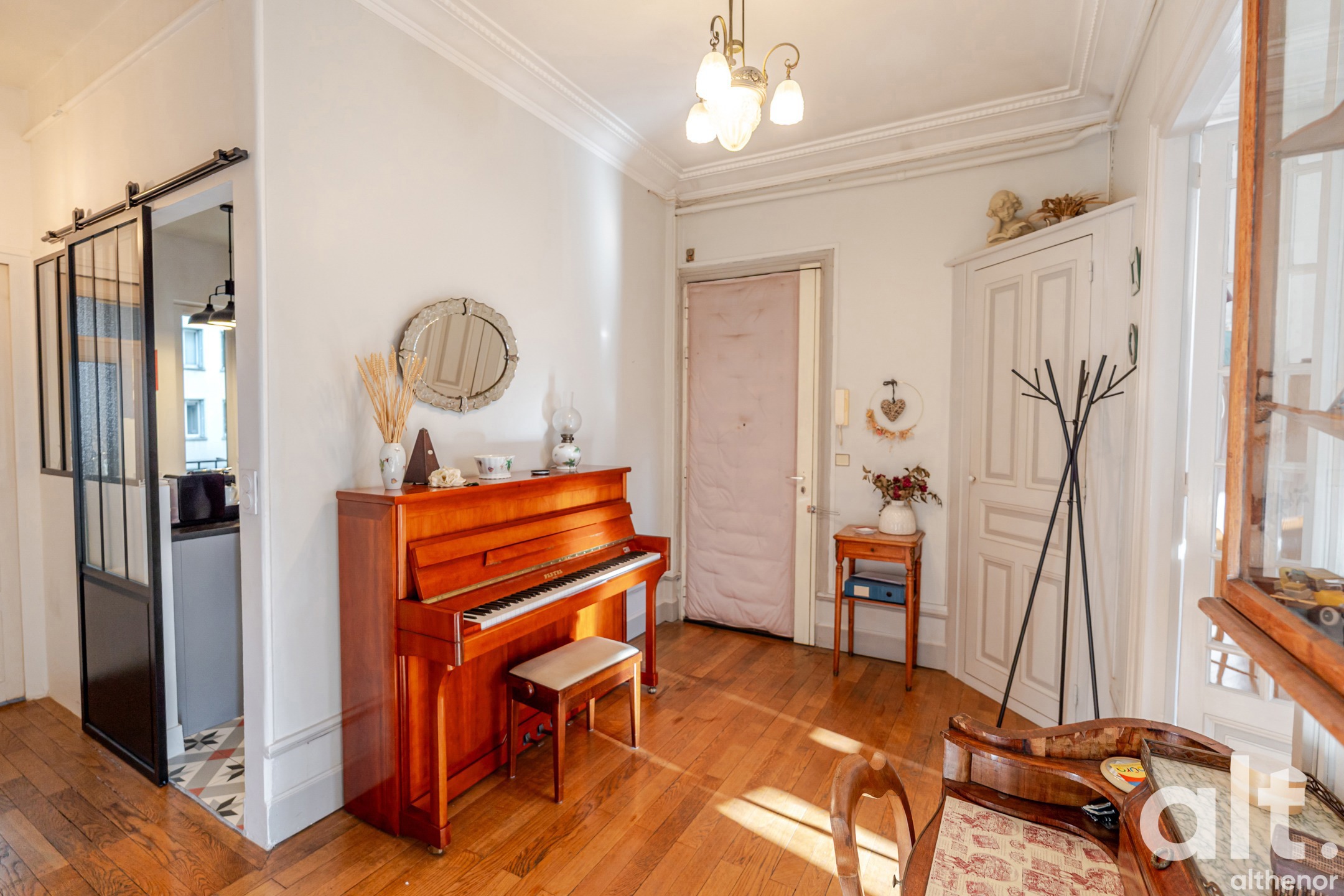 Spacious interior with a red wood upright piano, white doors, light parquet flooring, gilt chandelier, round mirror, small wooden table and scattered decorative accessories.