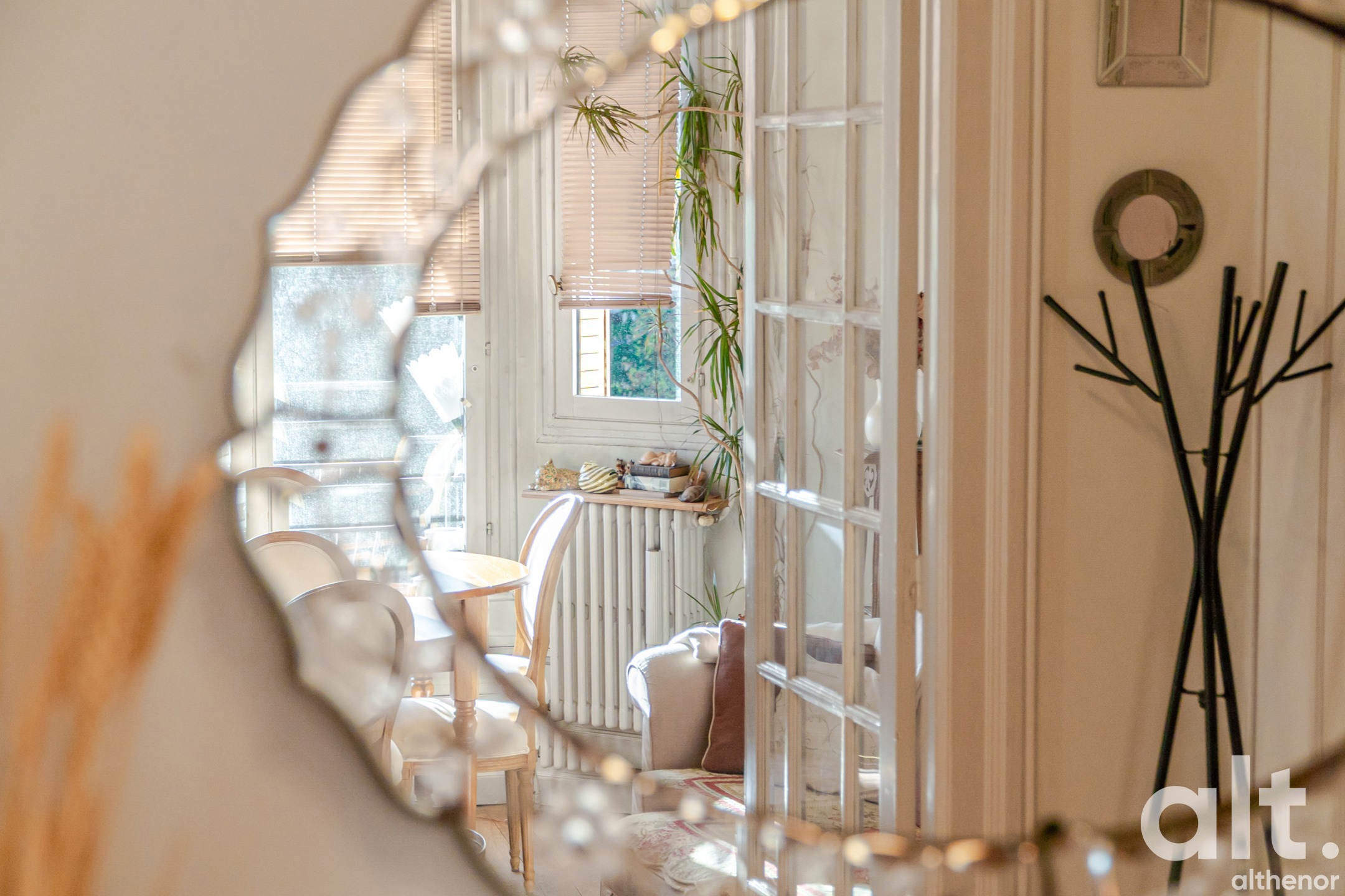Bright interior with open white glass doors leading to a room furnished with light-coloured chairs, hanging plants, radiator and wall decoration.