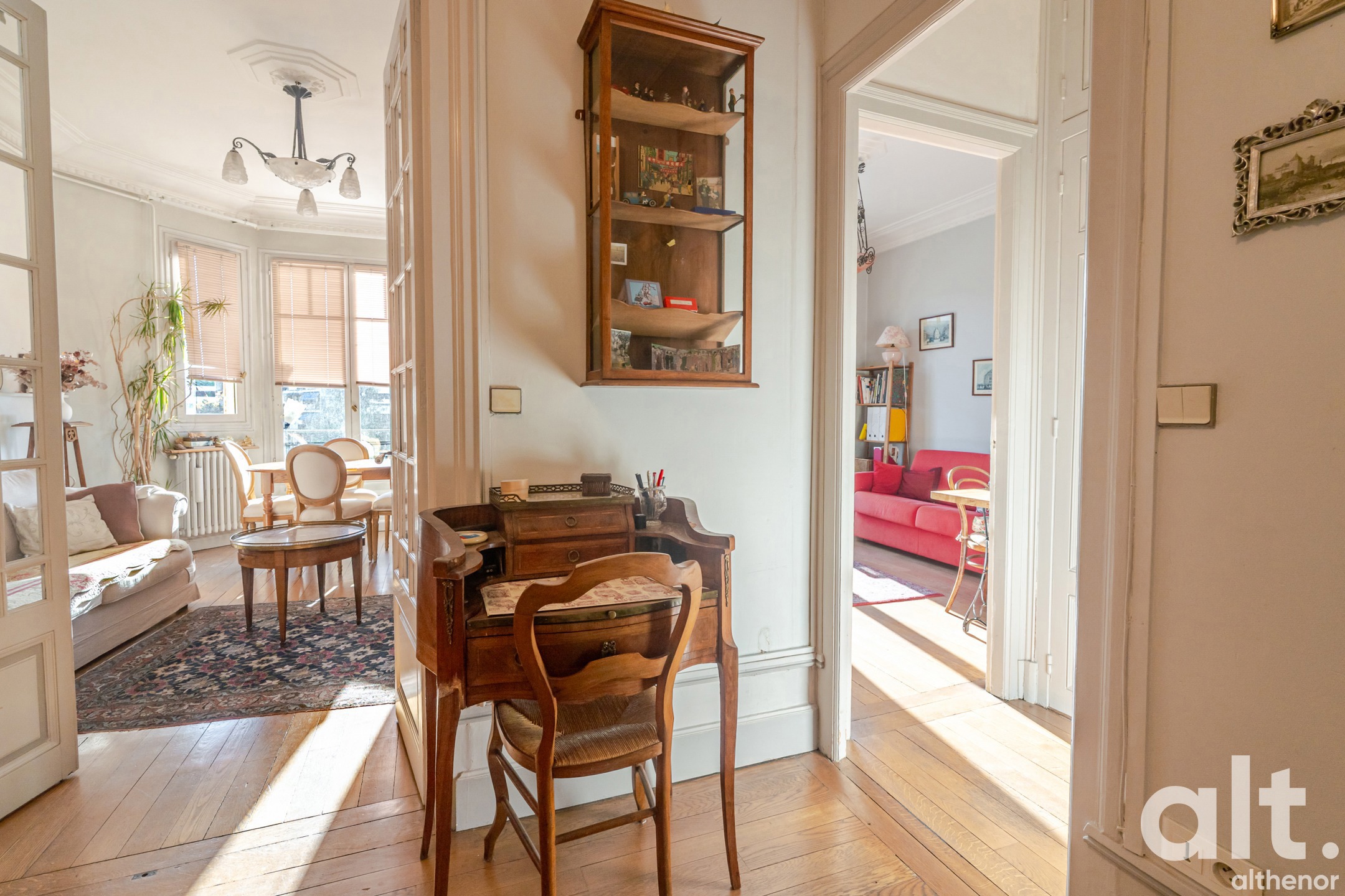 Bright interior with a wooden desk, matching chair, wall display cabinet, open doors leading to the living room and adjoining room, light parquet flooring and light beige walls.