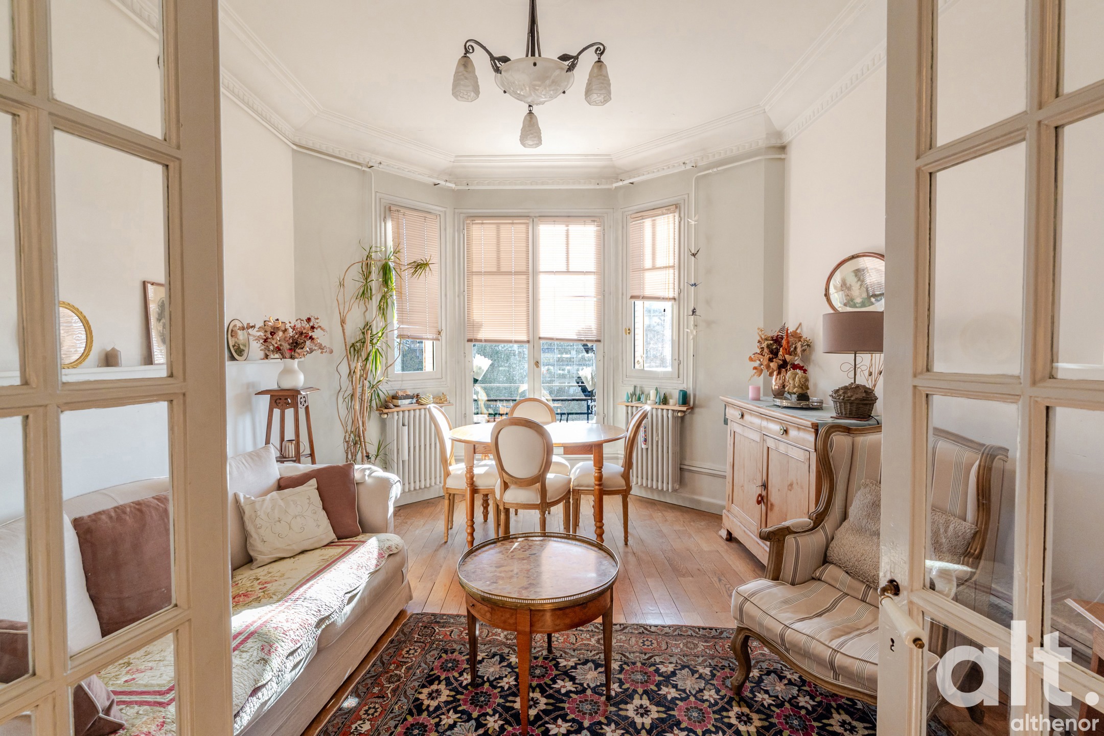 Bright living room with French windows opening onto a balcony, light wood furniture, beige sofa, central round table, hanging chandelier and floral decoration.