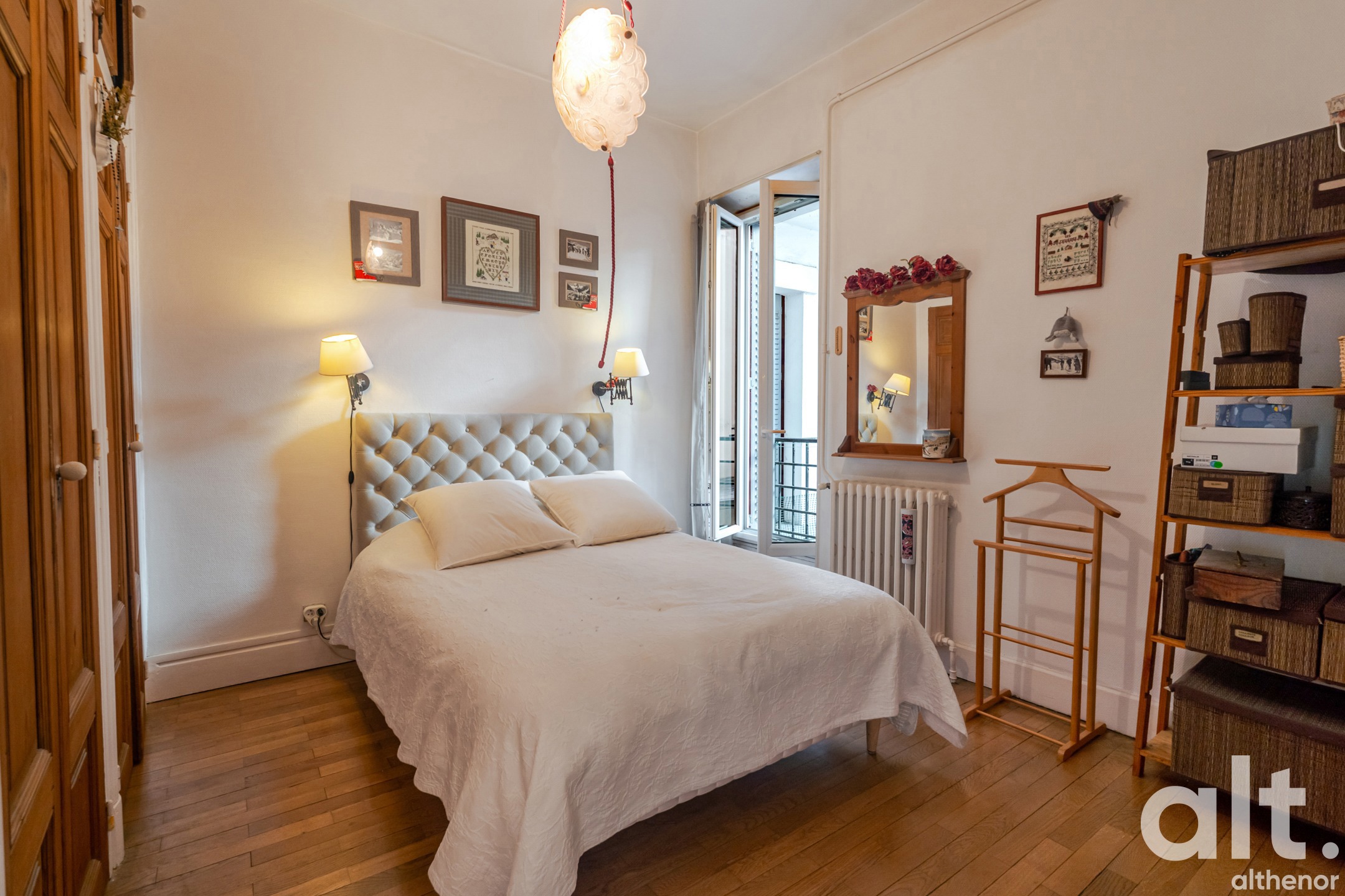 Bedroom with a double bed with a beige upholstered headboard, two bedside lamps, light parquet flooring, balcony accessible via French doors, mirror, wooden shelves and wicker storage.