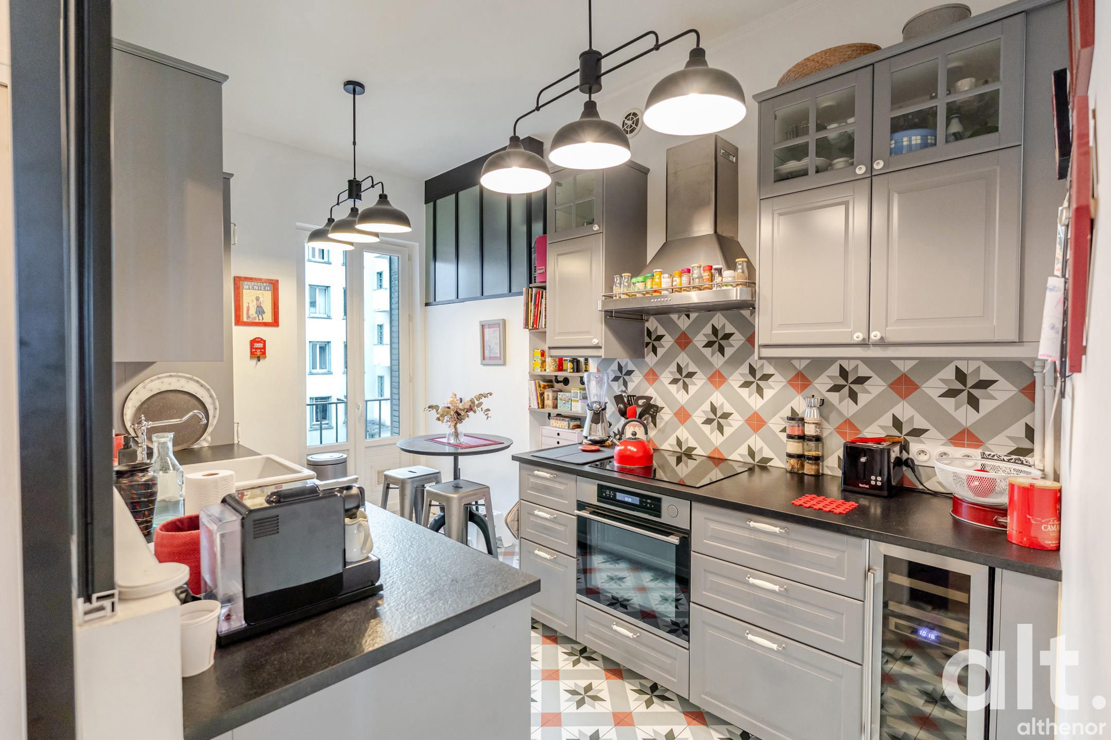 Fitted kitchen with grey units, black worktop, multicoloured geometric tiling, three metal pendant lights, window onto the courtyard, integrated appliances and access to the adjoining room.