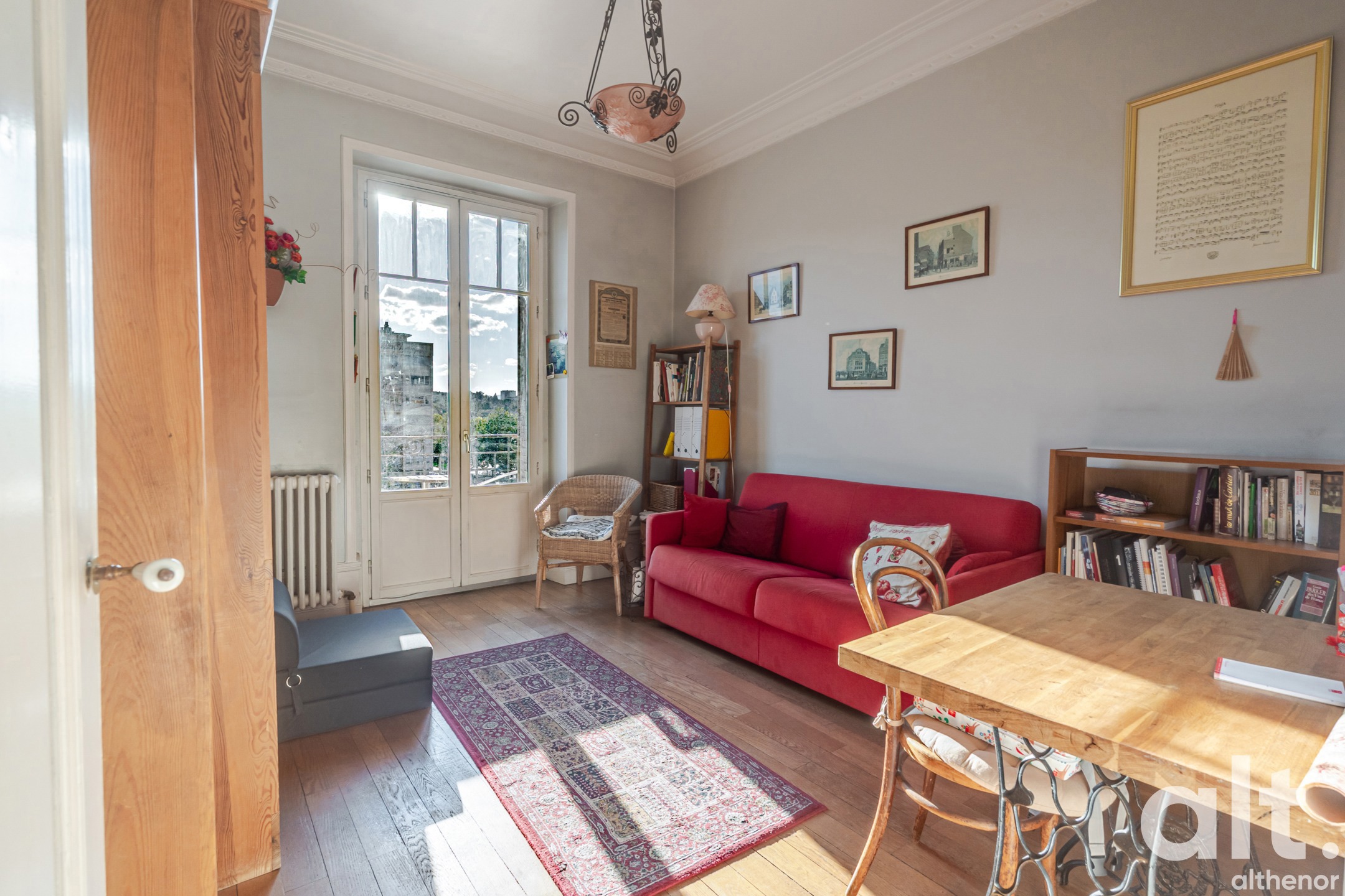 Bright living room with a red sofa, light wood table, bookcases, French doors opening onto a balcony, parquet flooring, wall frames and metal pendant light.