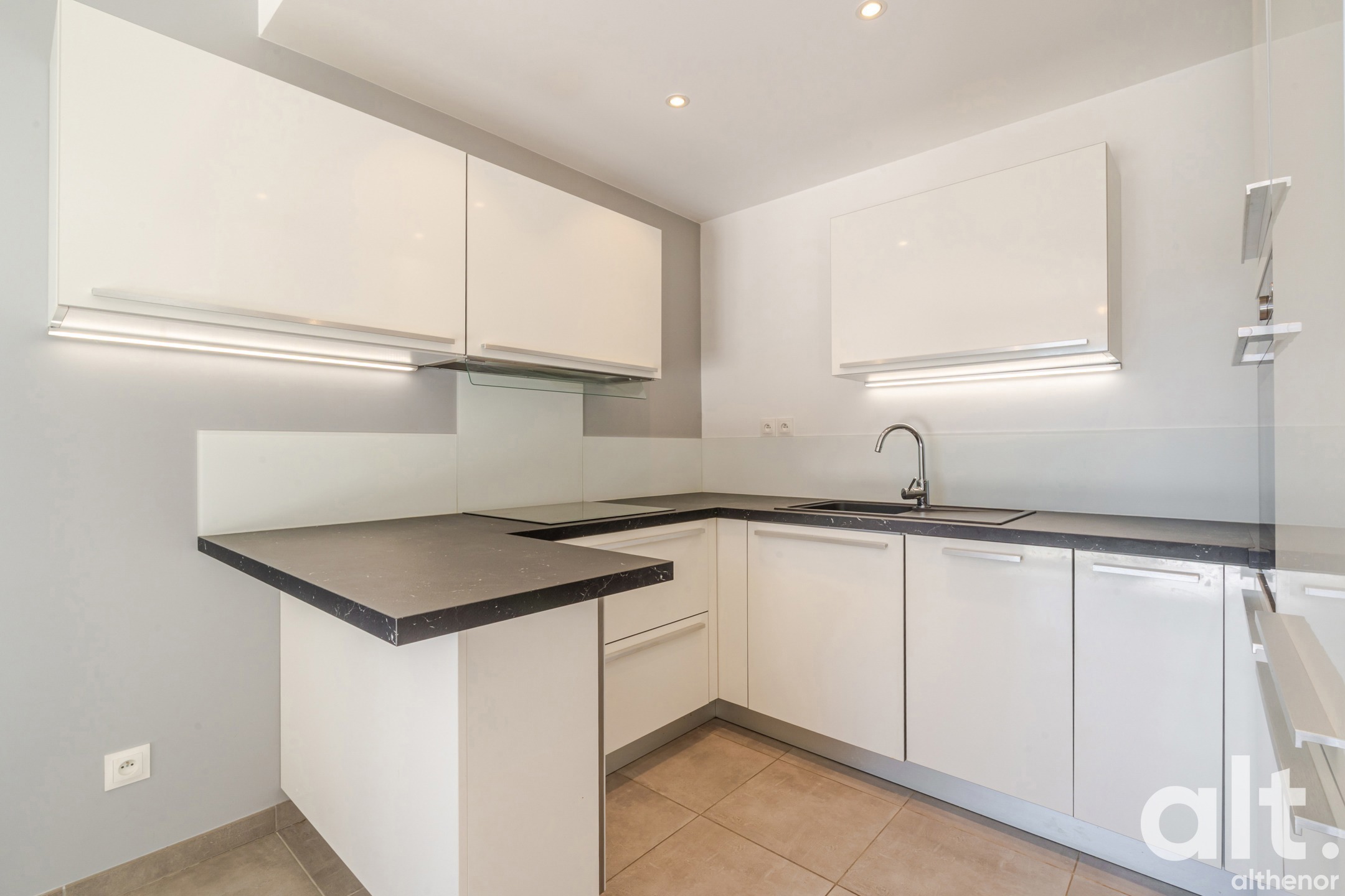 Modern kitchen with white units, black L-shaped worktop, chrome sink, illuminated wall cabinets and terracotta floor tiling.