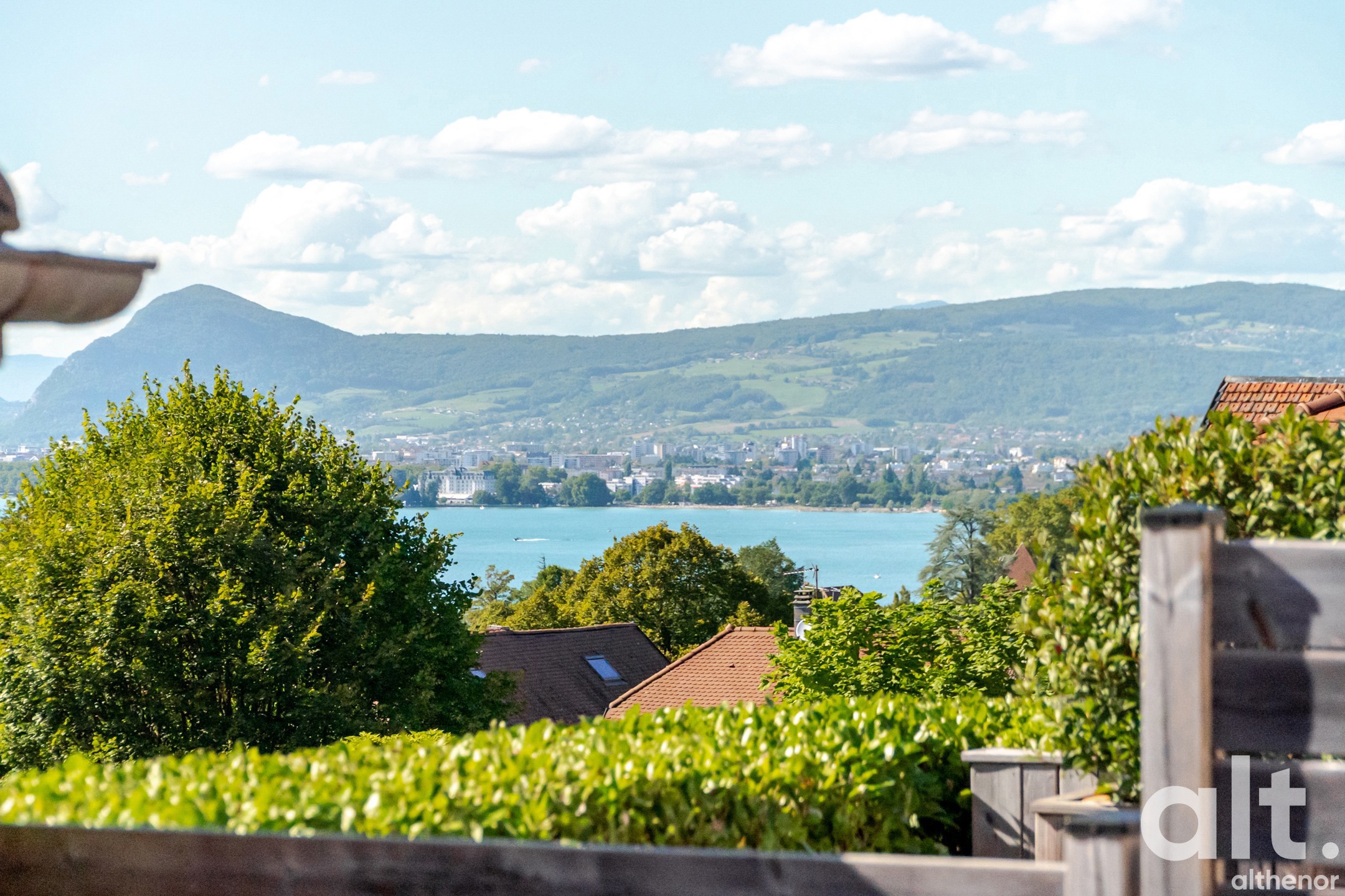 Extérieur, vue depuis une terrasse sur un lac bleu entouré de montagnes boisées, maisons aux toits rouges en premier plan, végétation dense.