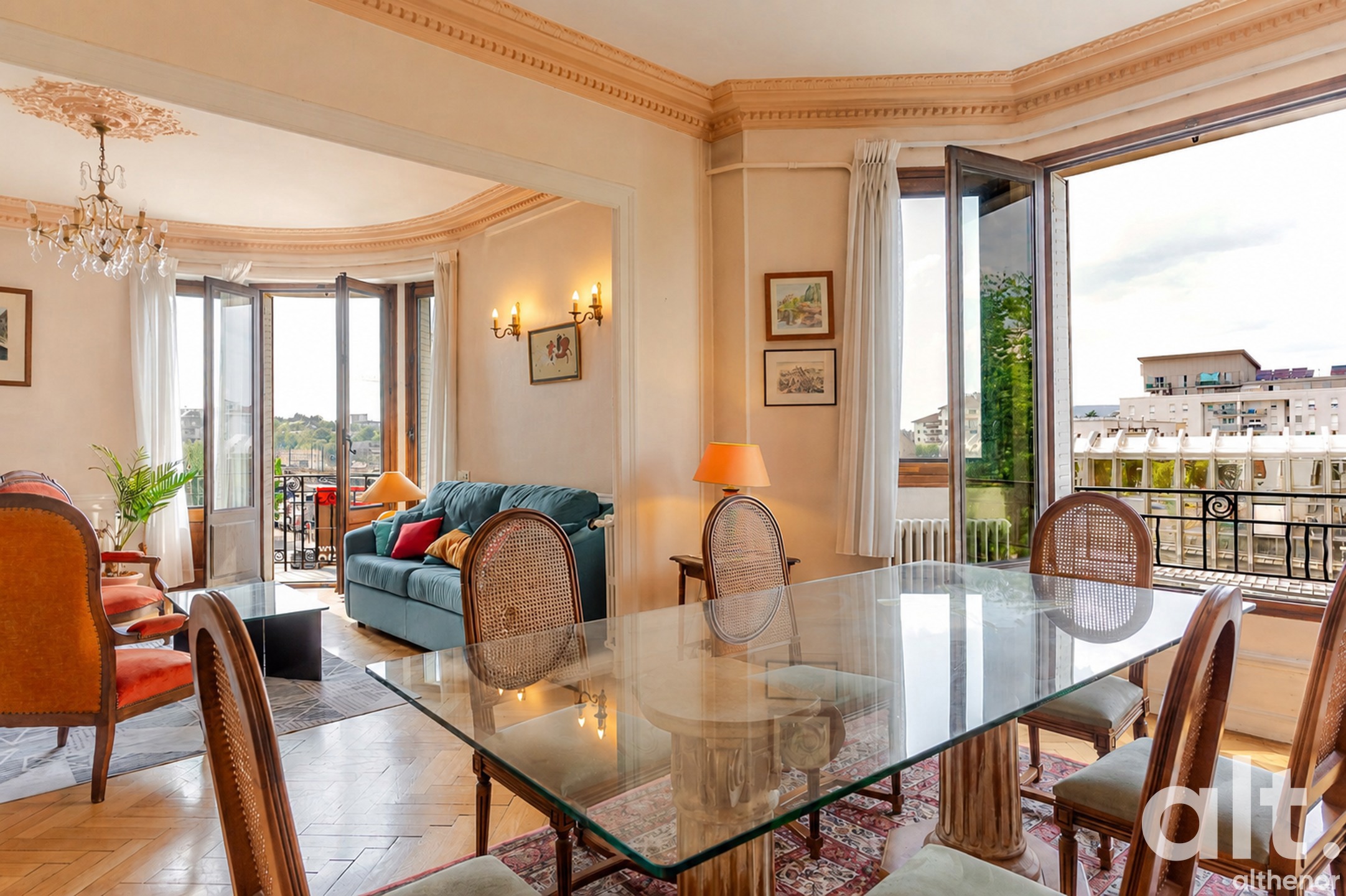 Intérieur d'appartement haussmannien avec salle à manger au premier plan, table en verre et chaises cannées, salon visible au fond avec canapé bleu, baies vitrées donnant sur balcon et vue urbaine.