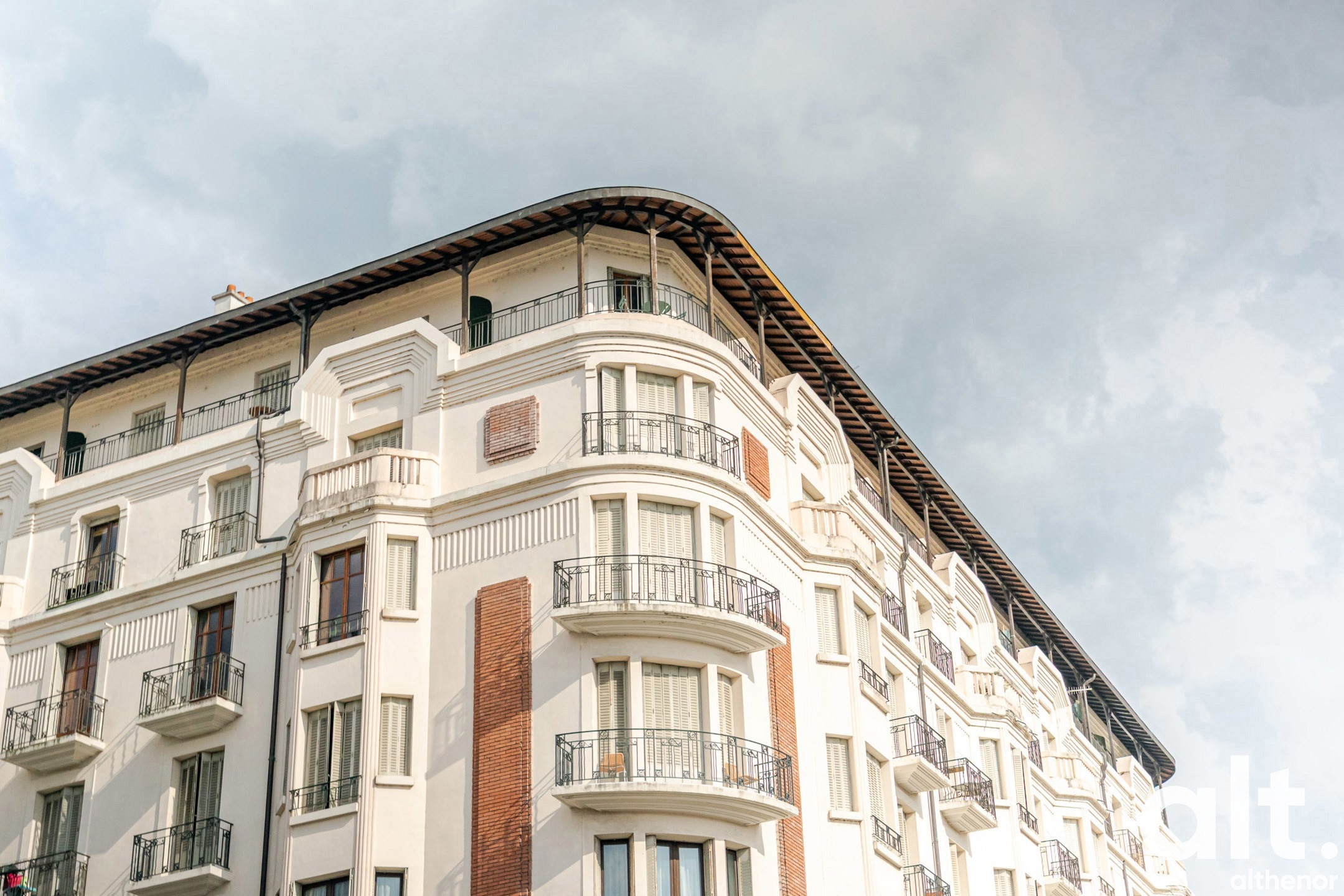 Immeuble d'angle de style haussmannien avec façade blanche, balcons courbes, fenêtres régulières et détails architecturaux en pierre, toiture mansardée.