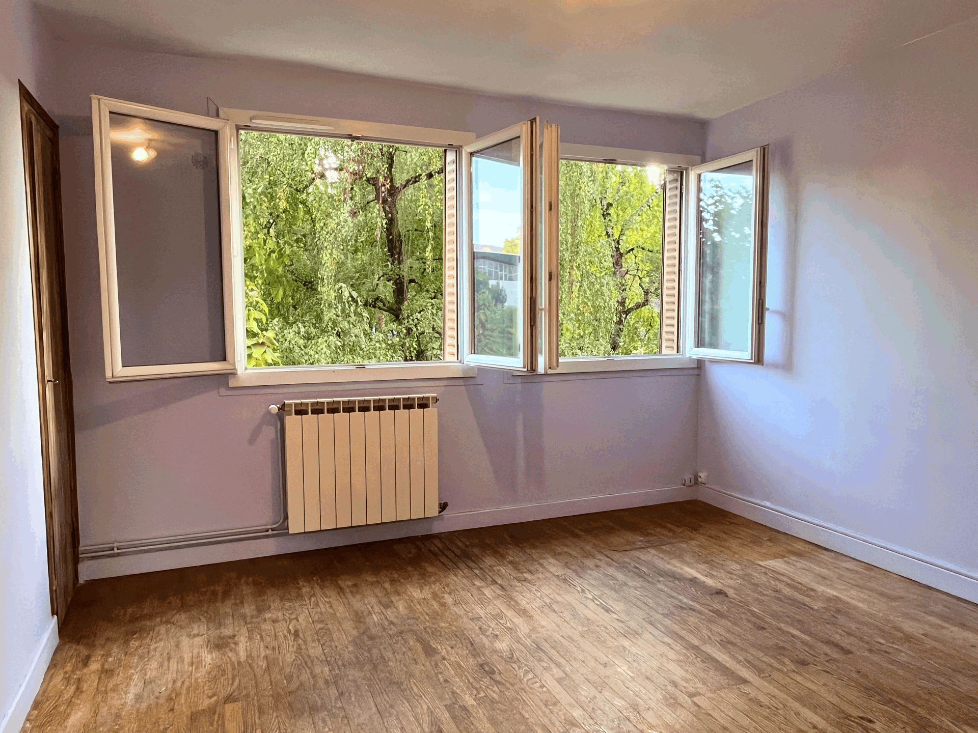 Interior of a room with lavender walls, light wood parquet floor, two open windows overlooking greenery, white metal radiator under the window, door on the left.